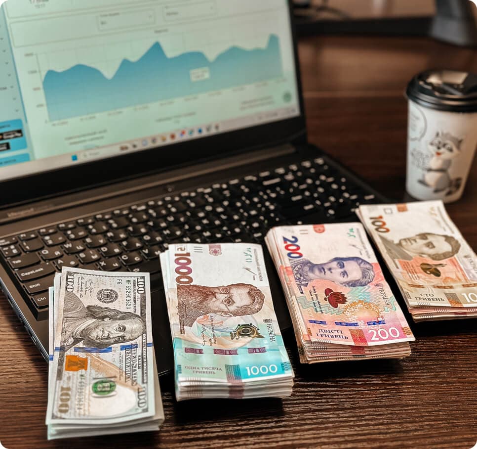 Ноутбук з відкритою програмою Kavapp, на якому лежать 4 пачки грошей у різній валюті, на фоні стаканчик з кавою від Coffee Mobile